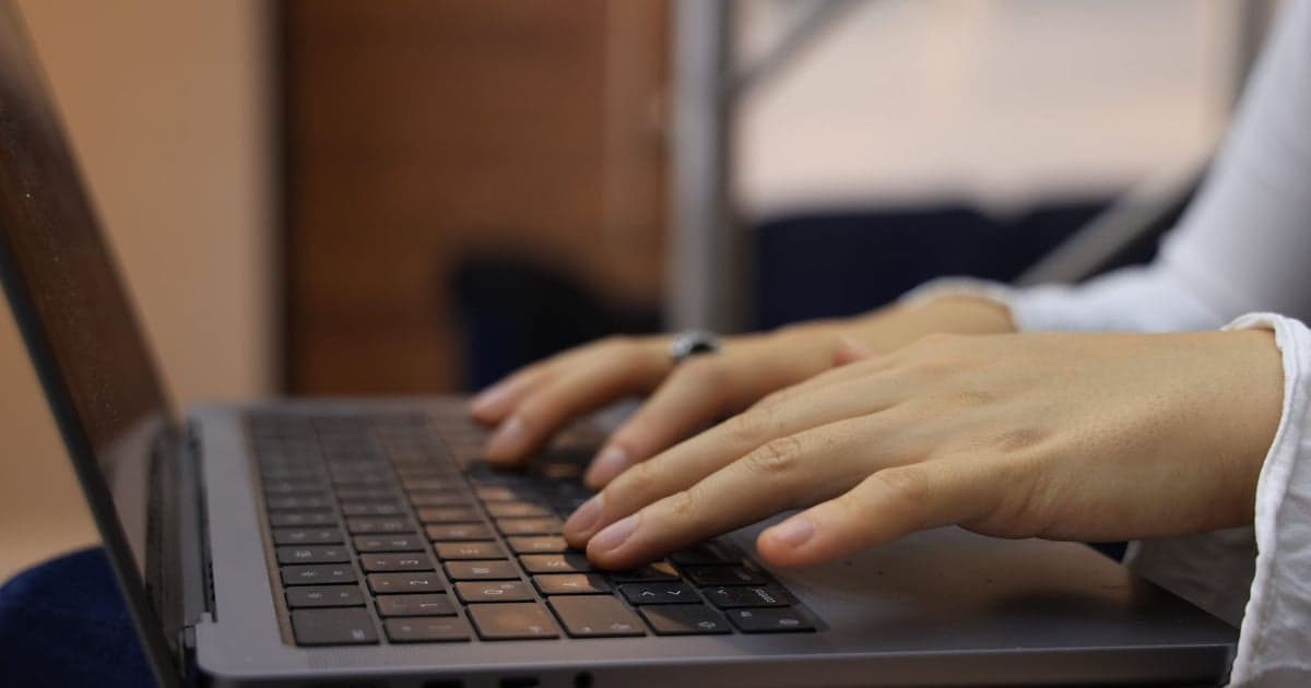 Mâinile unei persoane care scriu pe tastatura unui laptop, fotografiate din lateral